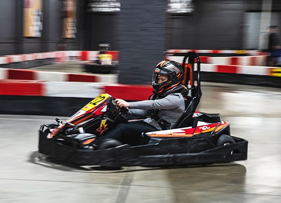 Image shows a person wearing a racing suit and helmet driving a go-kart on an indoor track.